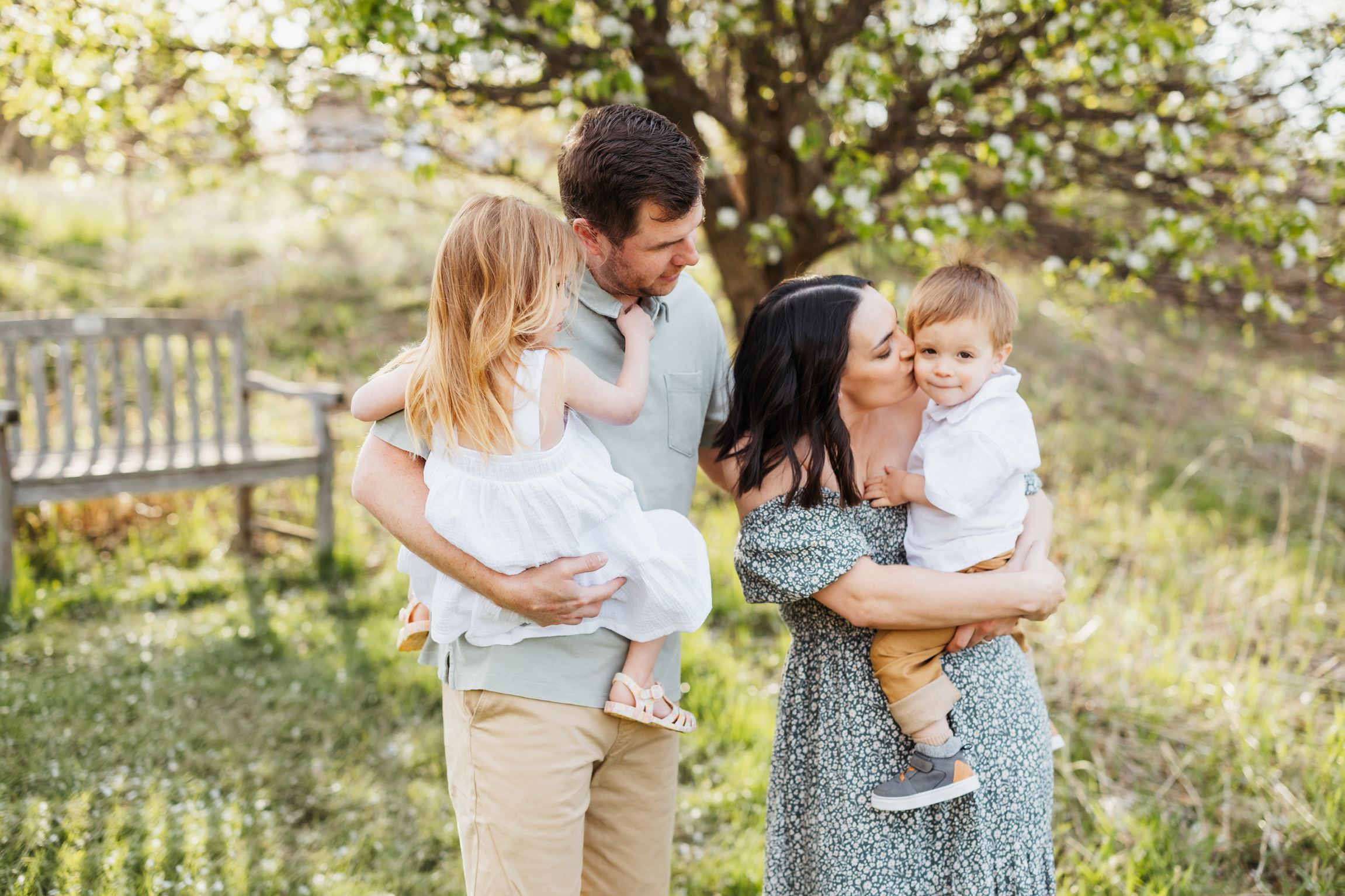 Family of four laughing together in a grassy park, each parent holding one of their children