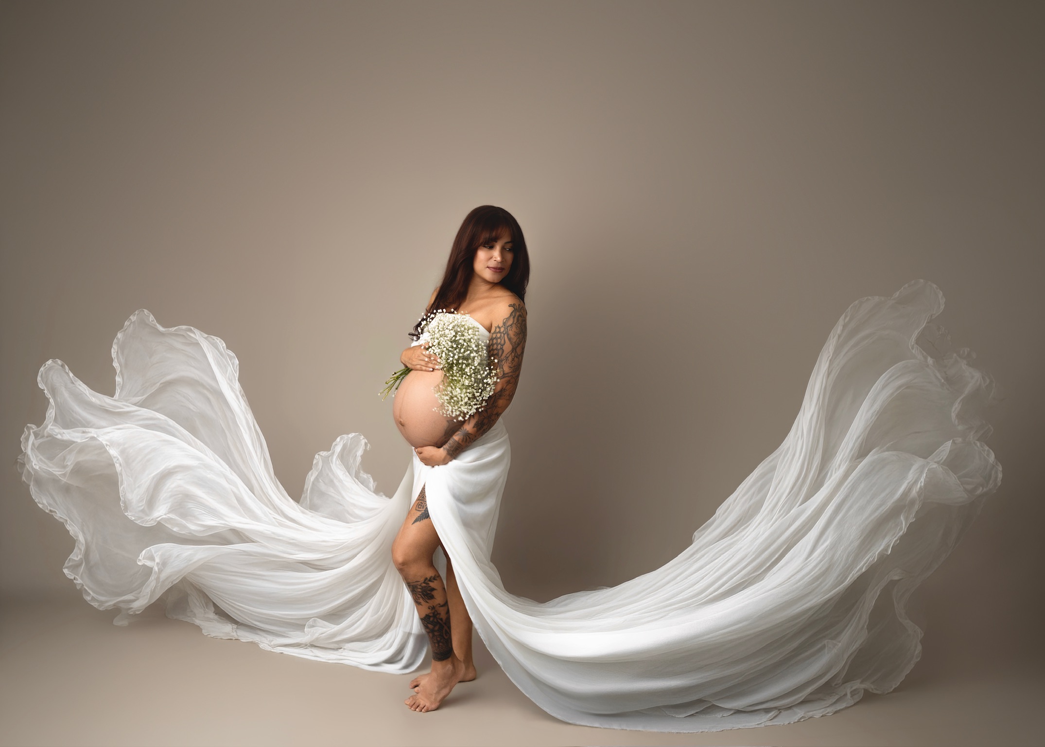 Expectant mother in a white gown holding a bouquet of baby's breath as long white fabric sweeps across a taupe studio floor