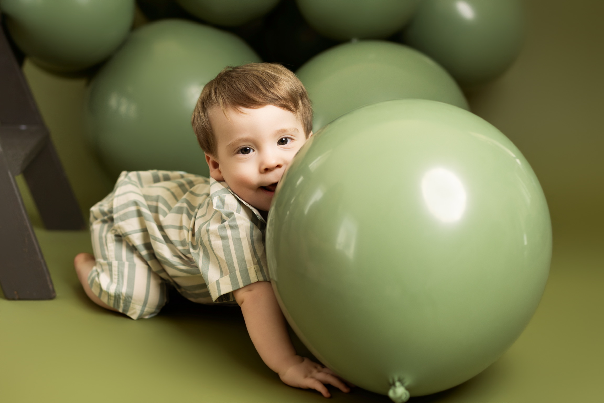 Toddler peeking over a large sage green balloon against an olive studio backdrop